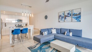 Un salon moderne avec un canapé d’angle bleu, une table basse blanche et un tapis bleu. On y trouve également un coin cuisine avec des meubles blancs et un tabouret de bar.