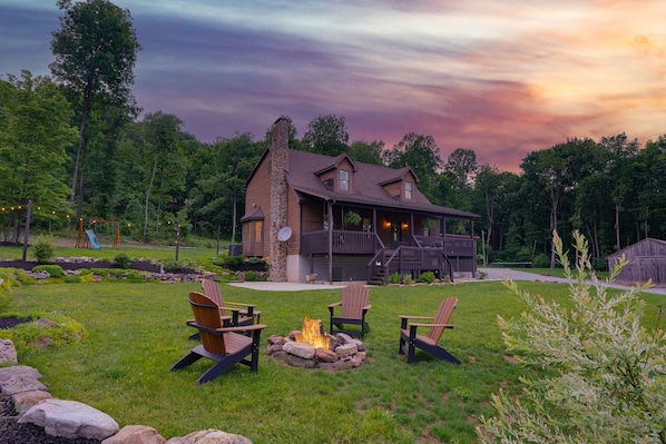 A house with a porch, a fire pit, and three Adirondack chairs.