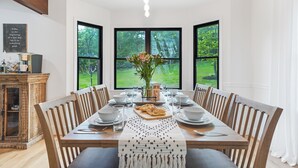 Comedor con una gran mesa dispuesta para seis personas, rodeada de sillas de madera. En la mesa hay un jarrón con flores, y un aparador de madera con puertas de cristal a la izquierda.