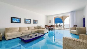 A living room with a beige sofa, a blue coffee table, wicker chairs, and a dining area with a view of the sea. - Case Marcia Mia - 2 adjacent seafront houses (San Montano)