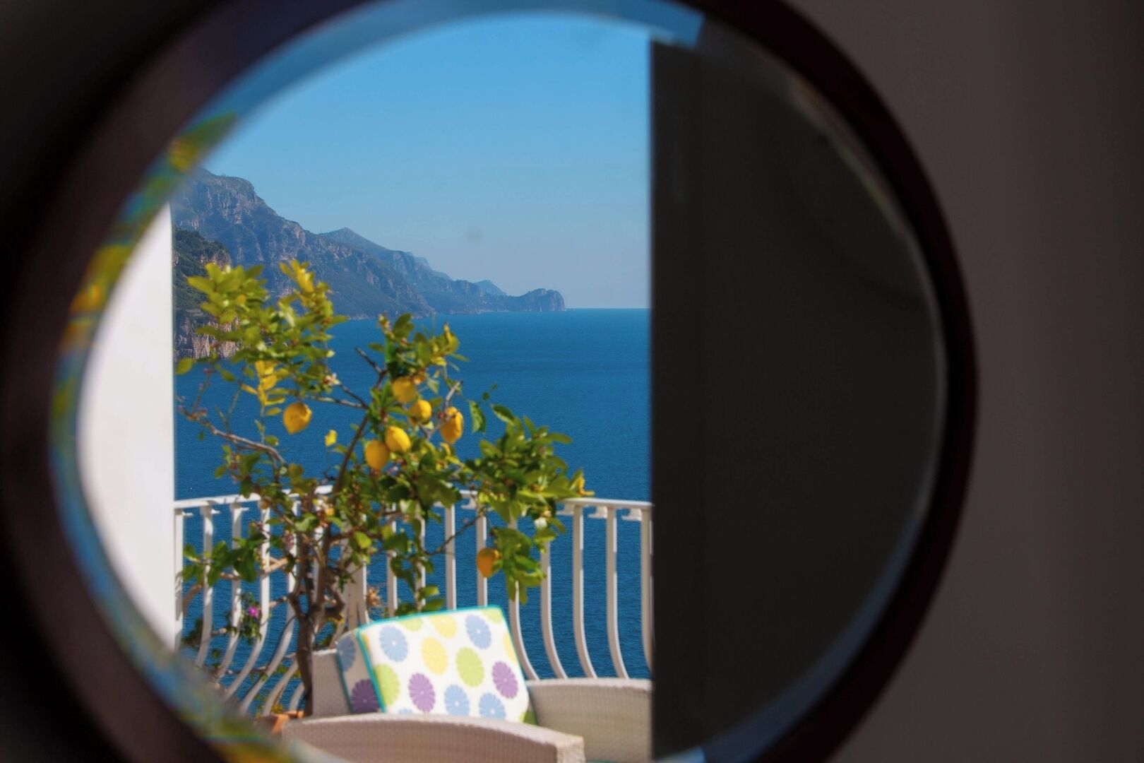 Une vue à travers une fenêtre donnant sur un balcon, avec une montagne et la mer en toile de fond.