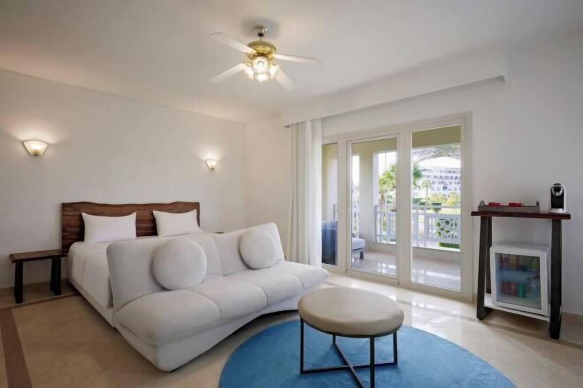 A modern bedroom with a white sofa, a round ottoman, a ceiling fan, and a bookshelf.