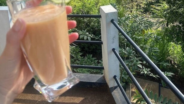 A hand holding a glass of iced coffee with a slice of lime, overlooking a lush valley with dense greenery and a clear sky. - Duet hearts (Ella)