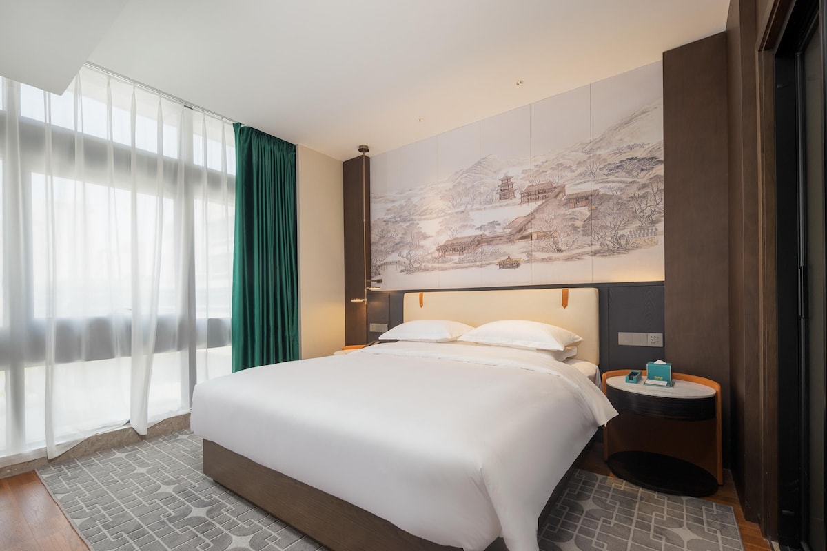 Habitación de hotel con una cama grande, una mesita de noche, un televisor montado en la pared y un ventanal con cortinas translúcidas.