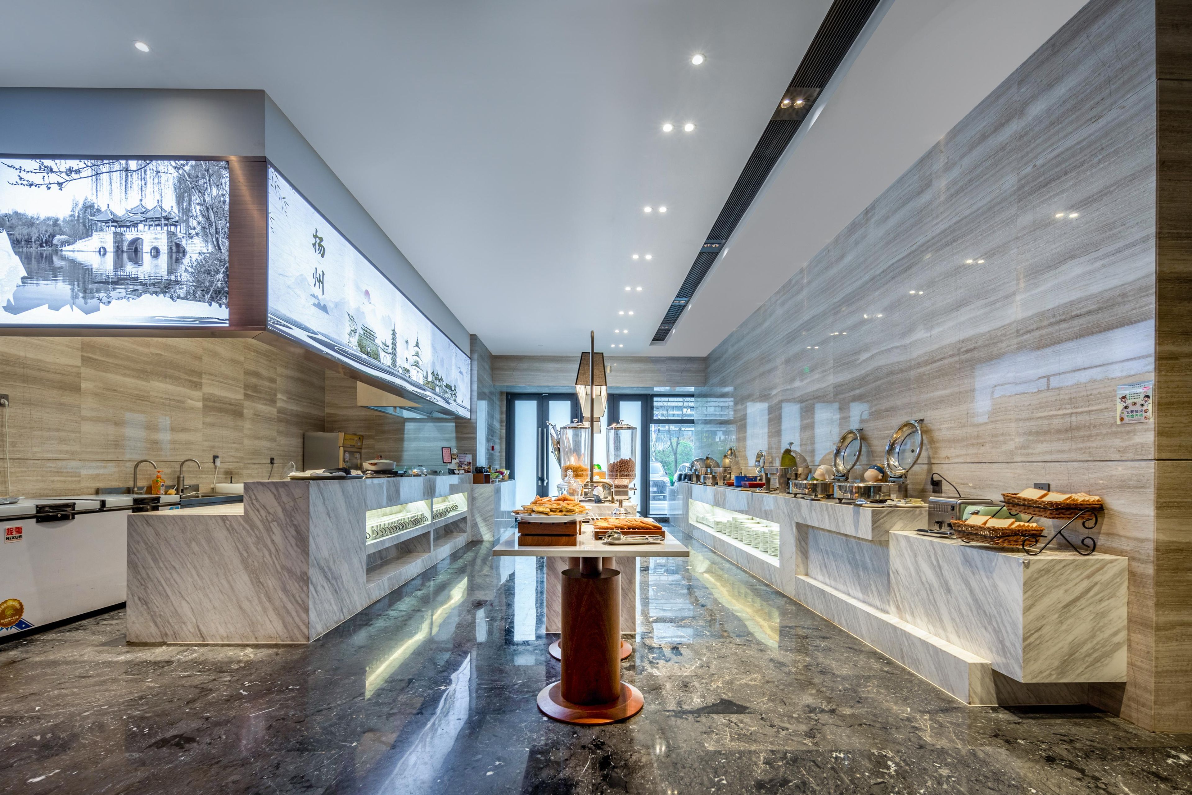 A modern hotel lobby with marble counters, a central chandelier, and a large digital screen displaying a scenic image.