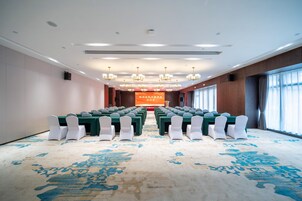 Un salón de conferencias con filas de mesas y sillas, un escenario con un cartel, y ventanas grandes con cortinas.