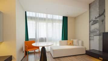 Quarto de hotel moderno com sofá, mesa de jantar e uma janela grande com cortinas.