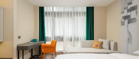 A hotel room with a bed, a desk, an orange chair, and a window with sheer curtains.