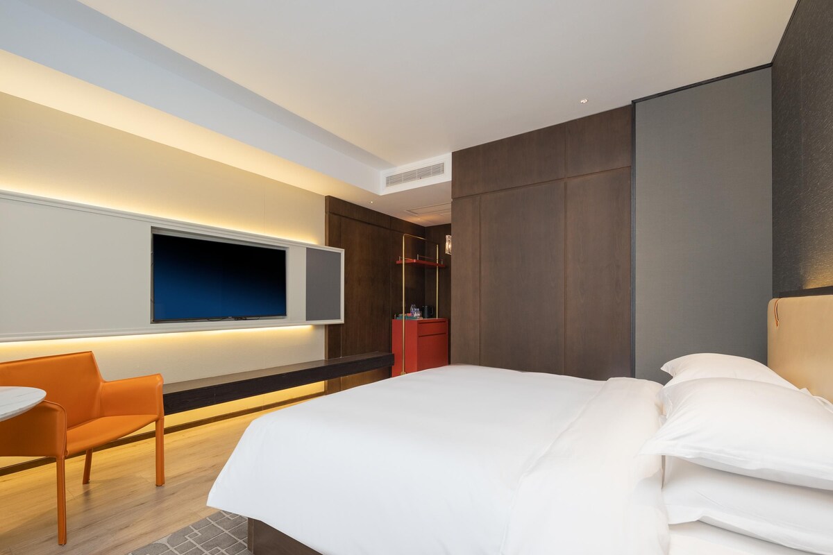 A modern hotel room with a large bed, a desk with a chair, a TV mounted on the wall, and a wardrobe.