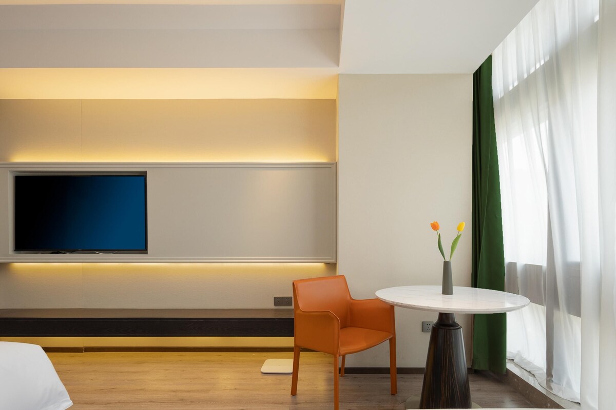 Una habitación de hotel moderna con un televisor de pantalla plana montado en la pared, una mesa redonda con un florero, una silla naranja y un ventanal con cortinas translúcidas.