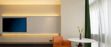 Een moderne hotelkamer met een flatscreen televisie aan de muur, een ronde tafel met een bloevas, een oranje fauteuil en een groot raam met dunne gordijnen.