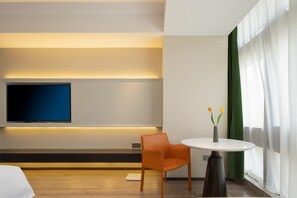 Una habitación de hotel moderna con un televisor de pantalla plana montado en la pared, una mesa redonda con un florero, una silla naranja y un ventanal con cortinas translúcidas.