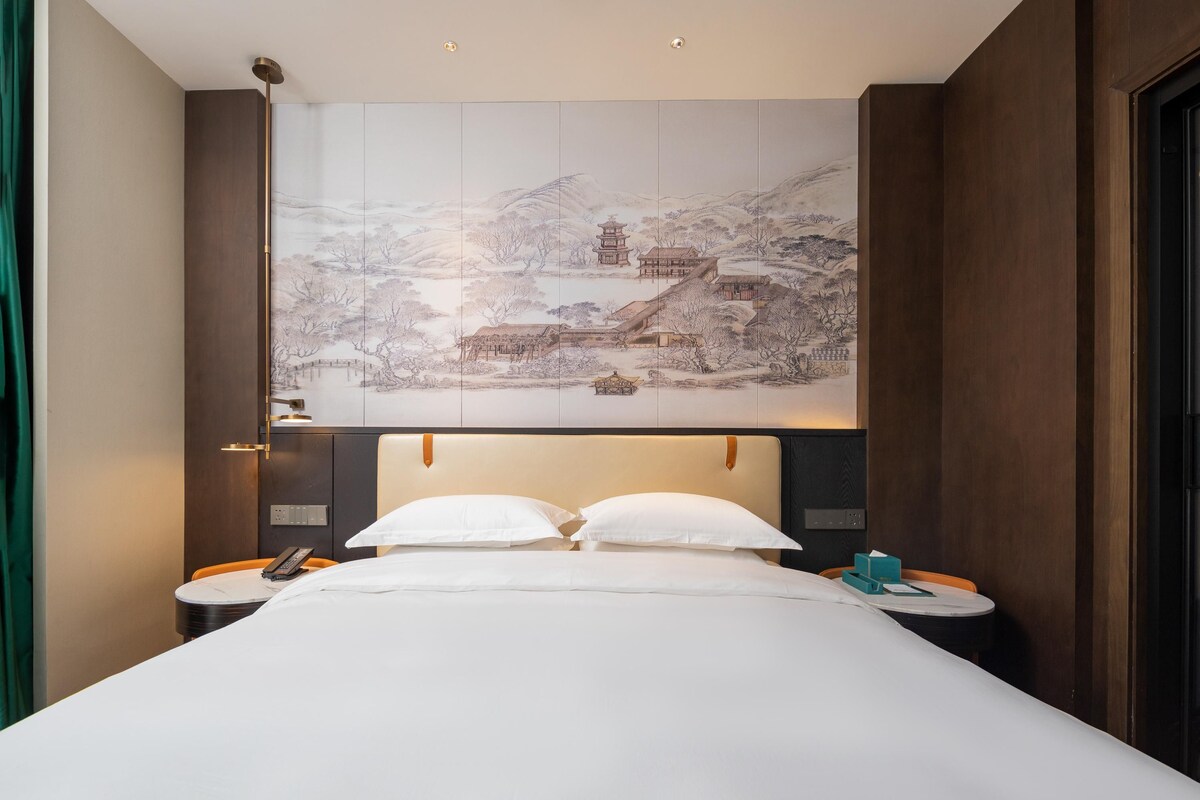 A hotel room with a large bed, two bedside tables, a wall-mounted artwork depicting a traditional scene, and a wooden headboard.