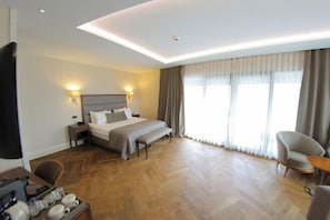 Une chambre d’hôtel comprenant un lit, des lampes de chevet, un bureau avec une chaise, une grande fenêtre avec des rideaux et un plancher en bois.