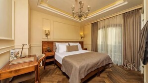 A hotel room with a large bed, a desk, a chair, a nightstand, a chandelier, and a large window with curtains. - Hotel Morione & Spa Center (Istanbul)