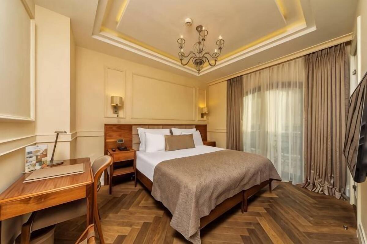 Una habitación de hotel con una cama grande, un escritorio, una silla, una mesita de noche, un candelabro y un amplio ventanal con cortinas.
