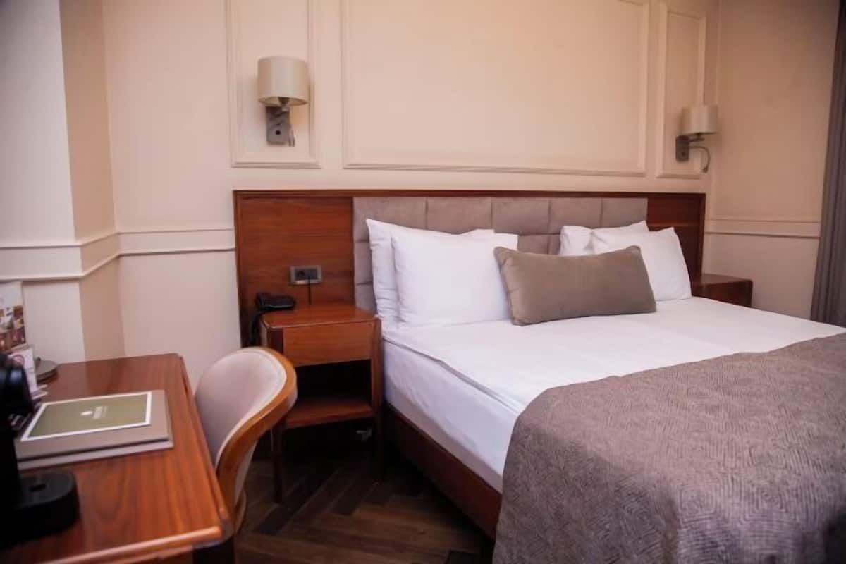 Una habitación de hotel con cama, mesita de noche, silla, lámpara y un pequeño escritorio con una computadora portátil.
