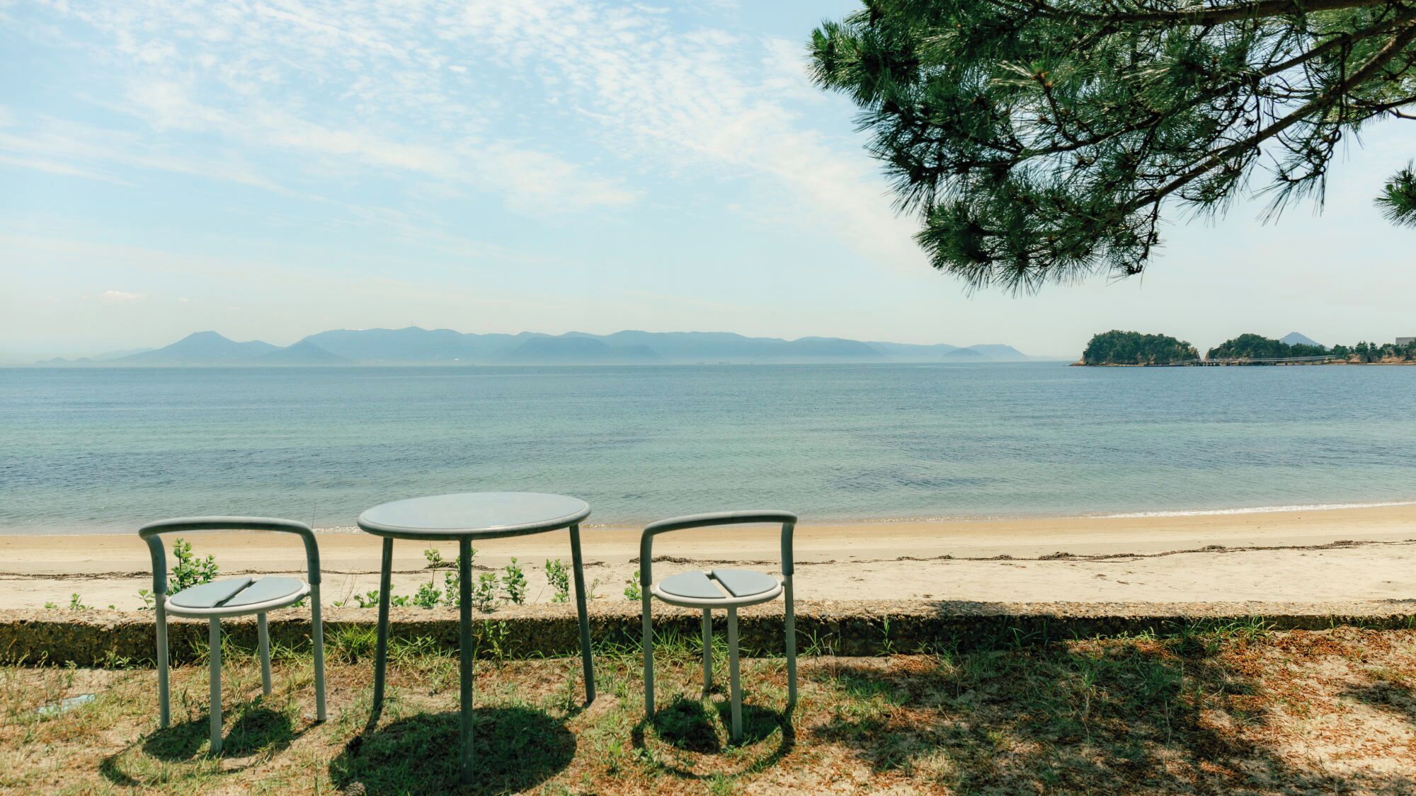 A beachside table with two chairs facing a calm sea, with distant mountains and a clear sky.