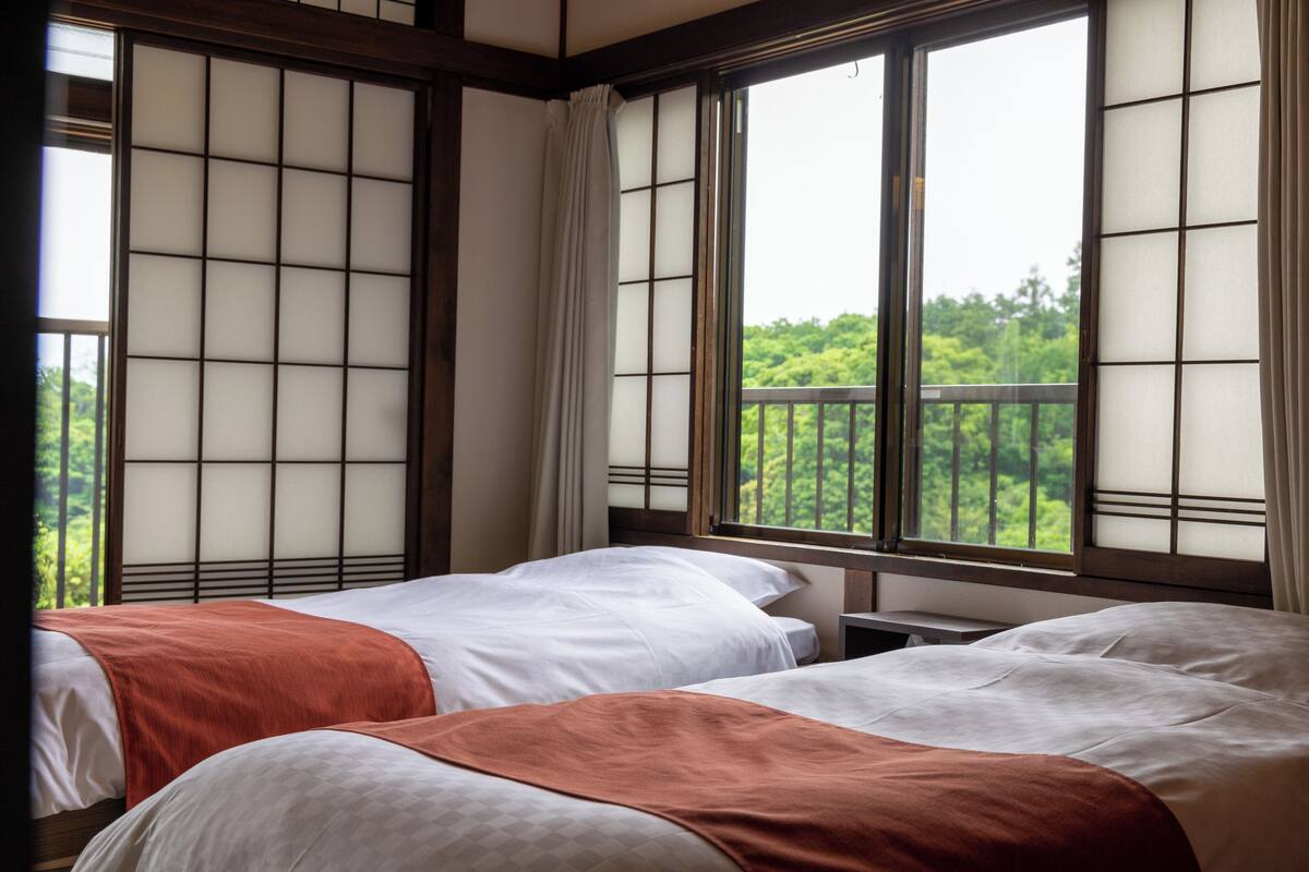 A room with two beds, large windows with a view of greenery, and a traditional Japanese-style sliding door.