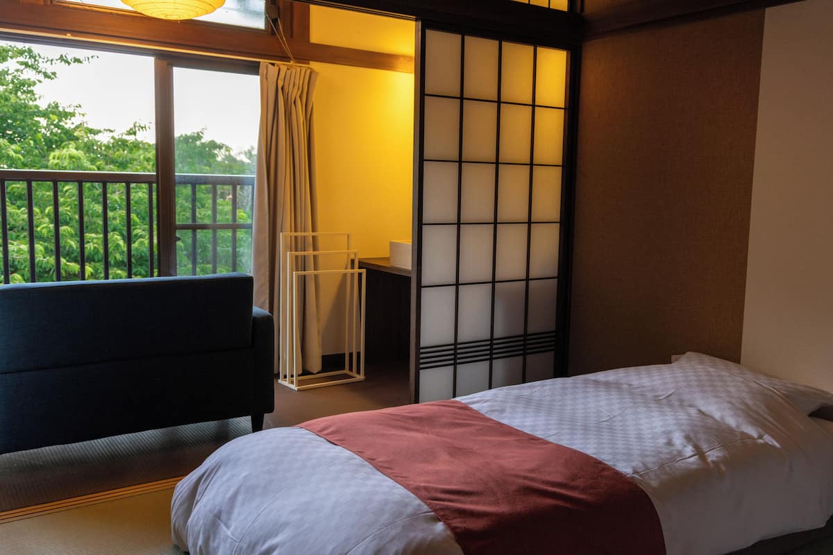 A hotel room with a bed, a sofa, a desk, and a view of greenery outside.