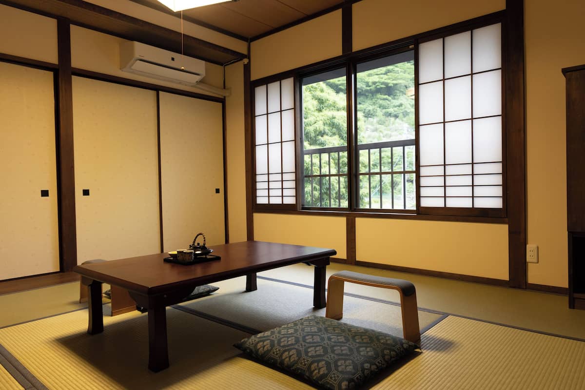 A traditional Japanese room with tatami flooring, a low table, a bench, and a large window with a view of greenery.