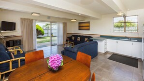 A modern living room with a sofa, dining table, and a TV. There is a kitchen area with cabinets and a window.