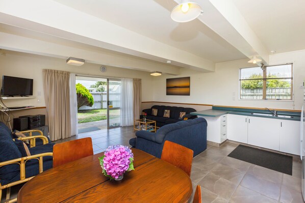 A modern living room with a sofa, dining table, and a TV. There is a kitchen area with cabinets and a window. - Paradise in Waikawa Beach - Family and pet friendly - Home away from Home (Waikawa Beach)