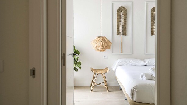 A bedroom with a bed, a hanging light fixture, a wall-mounted mirror, and a small stool.