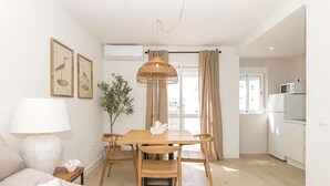 A modern kitchen with white appliances, a dining area with a wooden table and chairs, and a living room with a sofa and a lamp.