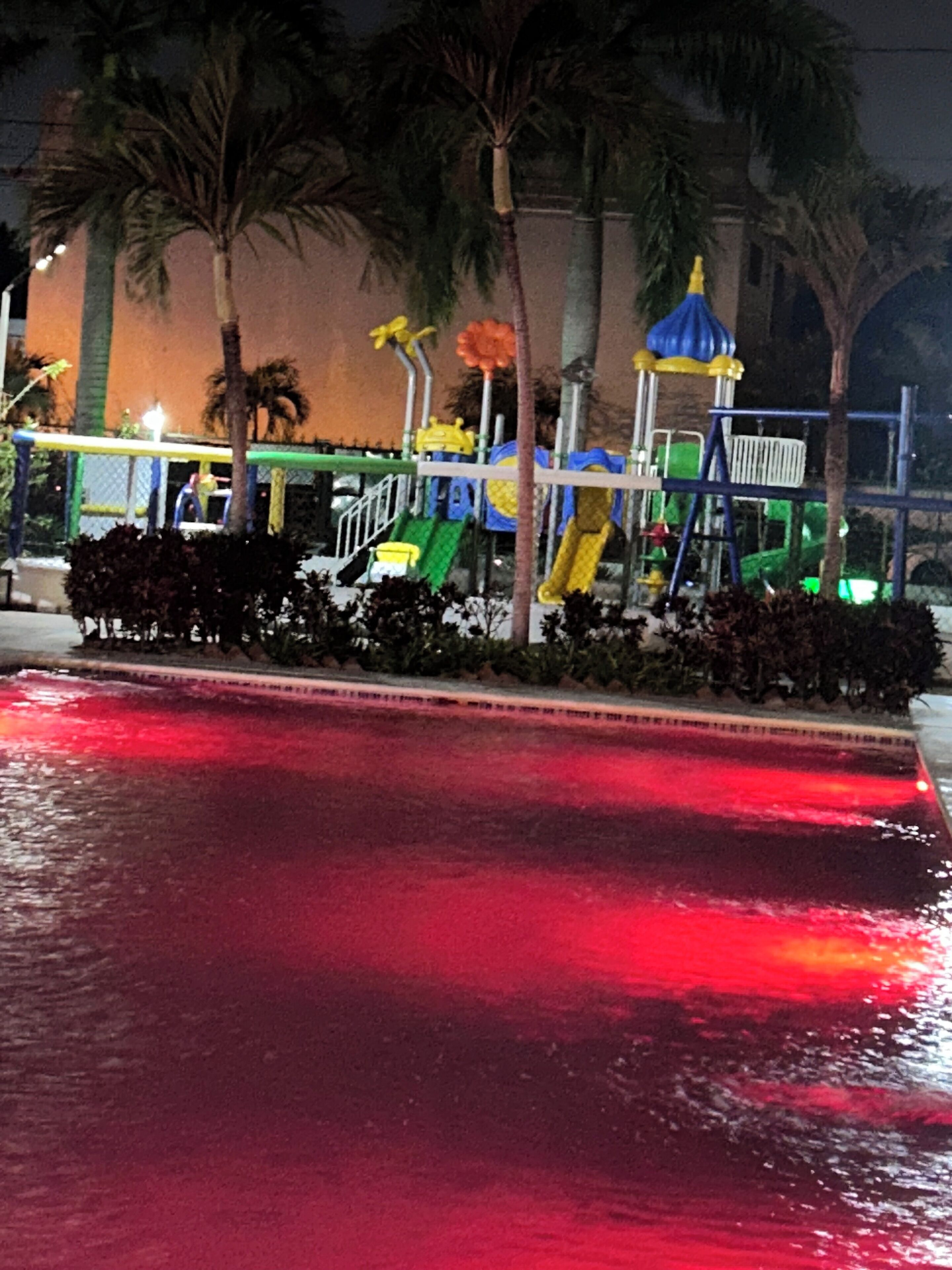 Vista noturna de um lago iluminado em vermelho, palmeiras e um parquinho com equipamentos coloridos.