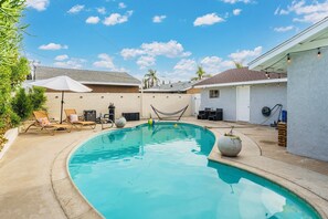 A swimming pool with lounge chairs and a hammock, surrounded by a fenced yard with a house and a clear sky.