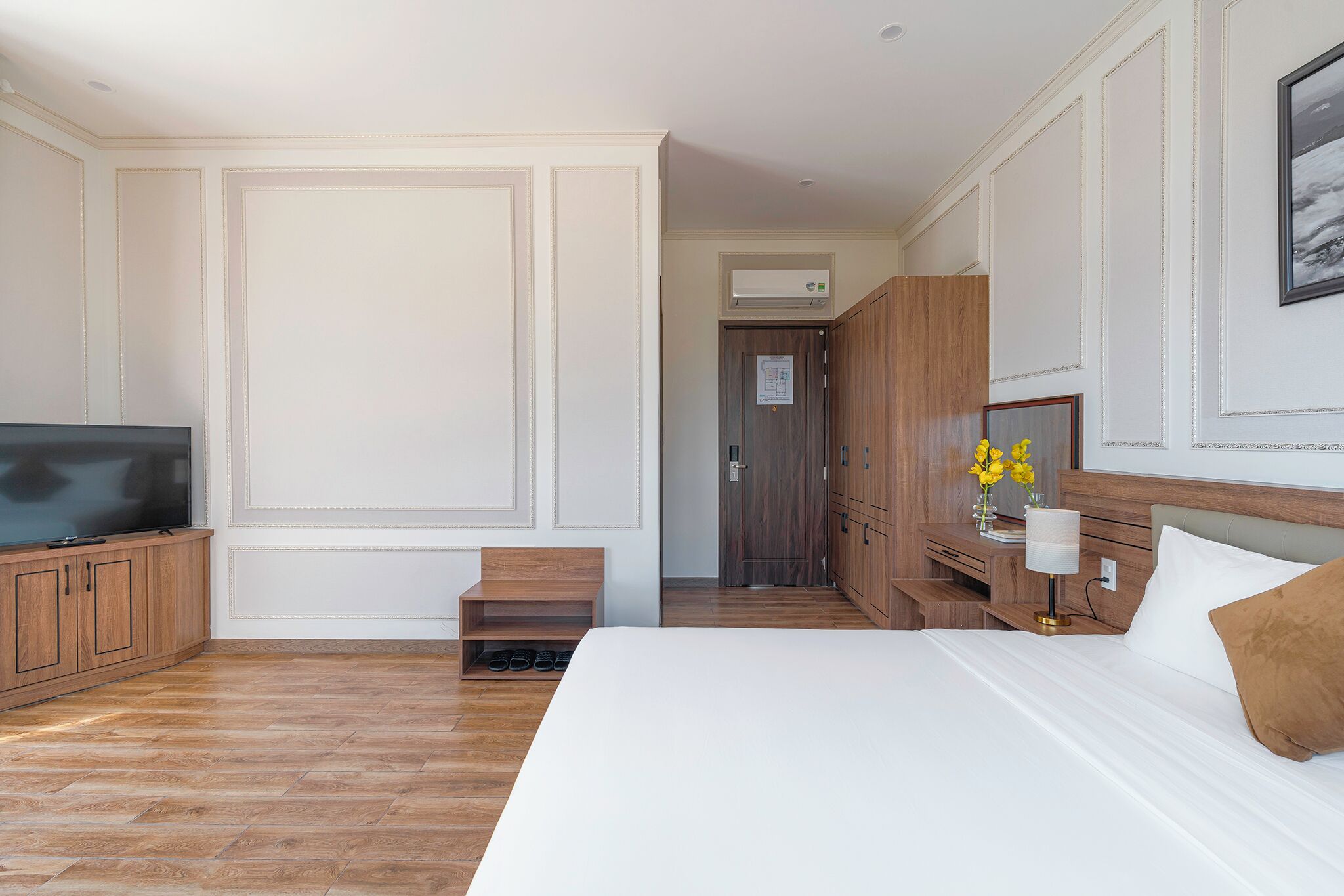 A hotel room with a large bed, wooden wardrobe, a TV, and a desk with a lamp and flowers.
