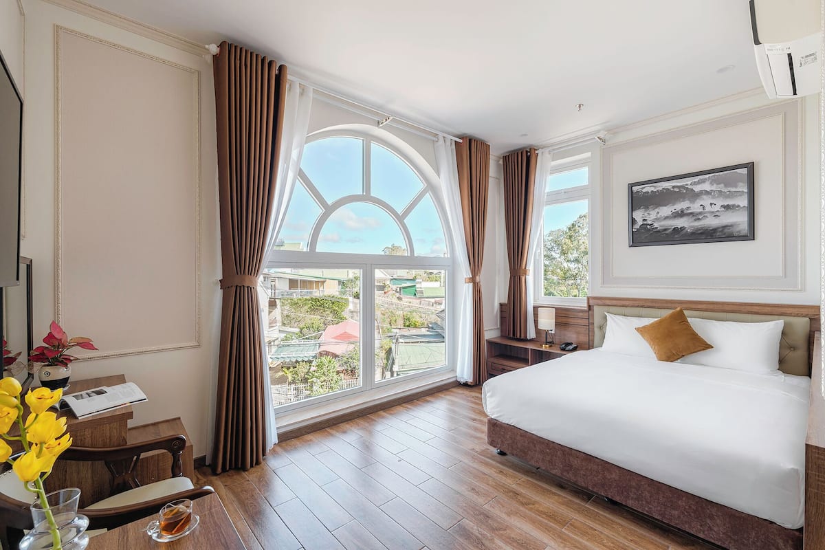 A hotel room with a large window, a bed, a desk with a chair, and a vase of flowers.