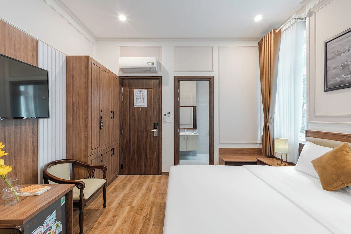 A hotel room with a wooden wardrobe, a desk with a chair, a bed with a white mattress and a brown pillow, a TV mounted on the wall, and a bathroom visible through a glass door.