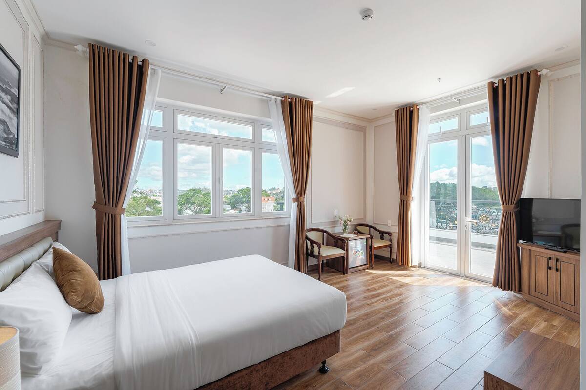 A hotel room with a large bed, a desk with chairs, a television, and large windows with curtains.