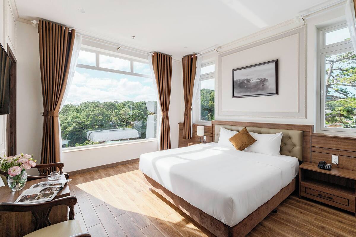 A hotel room with a large bed, wooden furniture, a desk with a vase of flowers, and large windows with curtains.