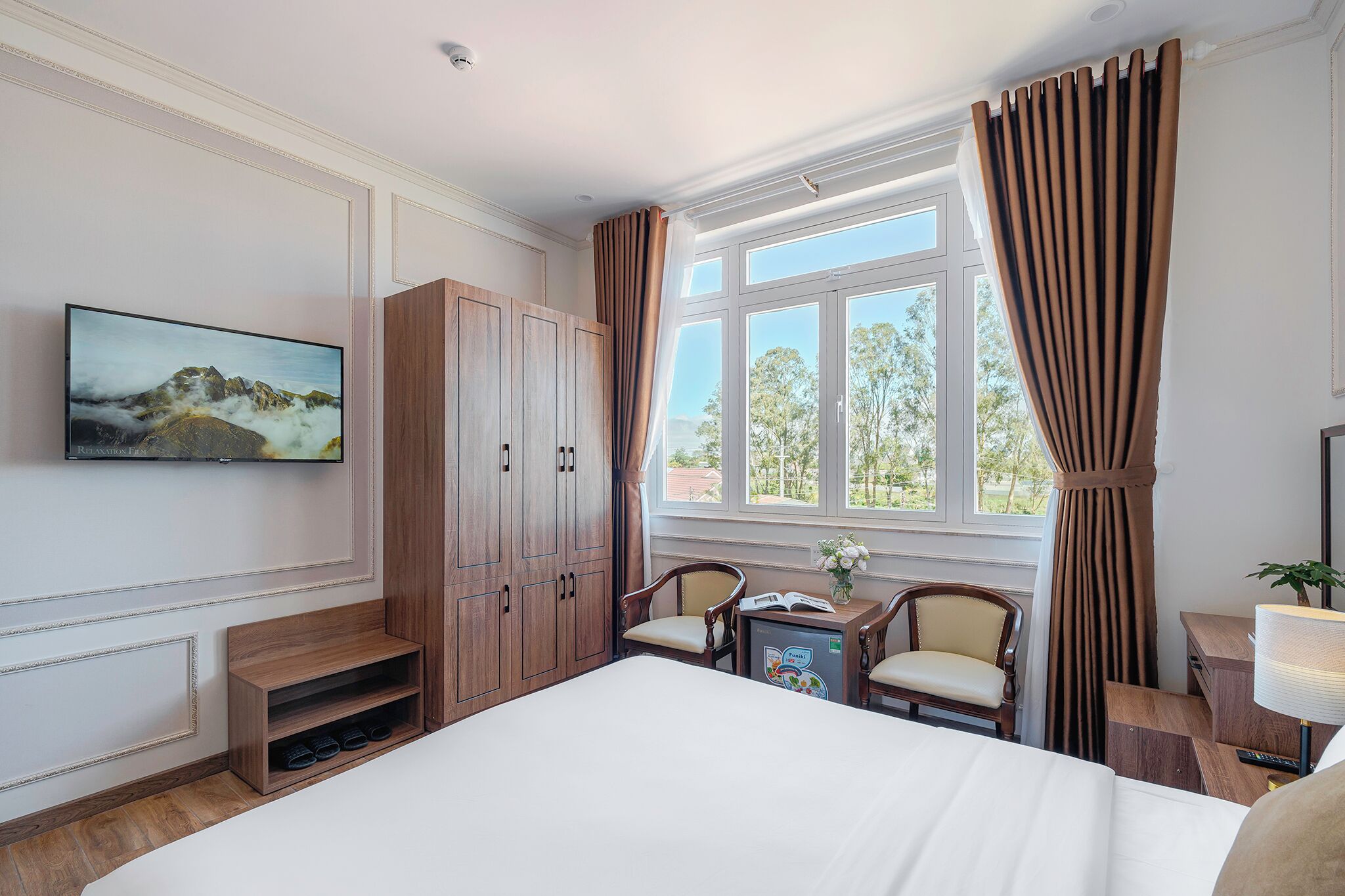 A hotel room with a large bed, wooden wardrobe, a desk with a chair, a small table with a lamp, and a TV mounted on the wall.