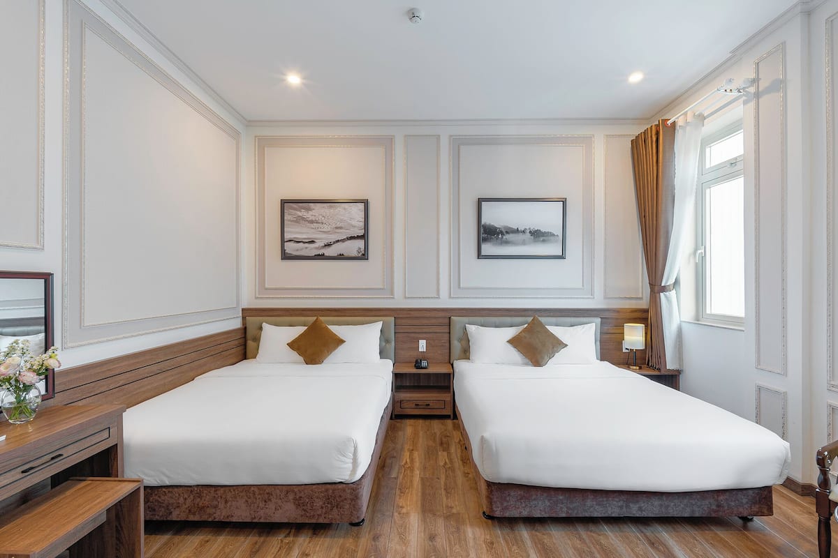 A hotel room with two beds, a nightstand with a lamp, a small table with a vase, and a window with curtains.
