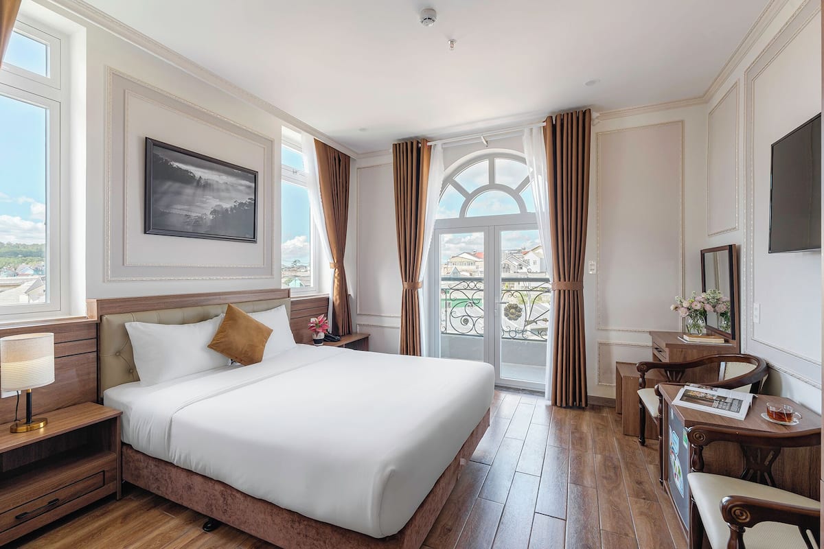 A hotel room with a large bed, a desk with a chair, a TV, and a balcony with curtains.