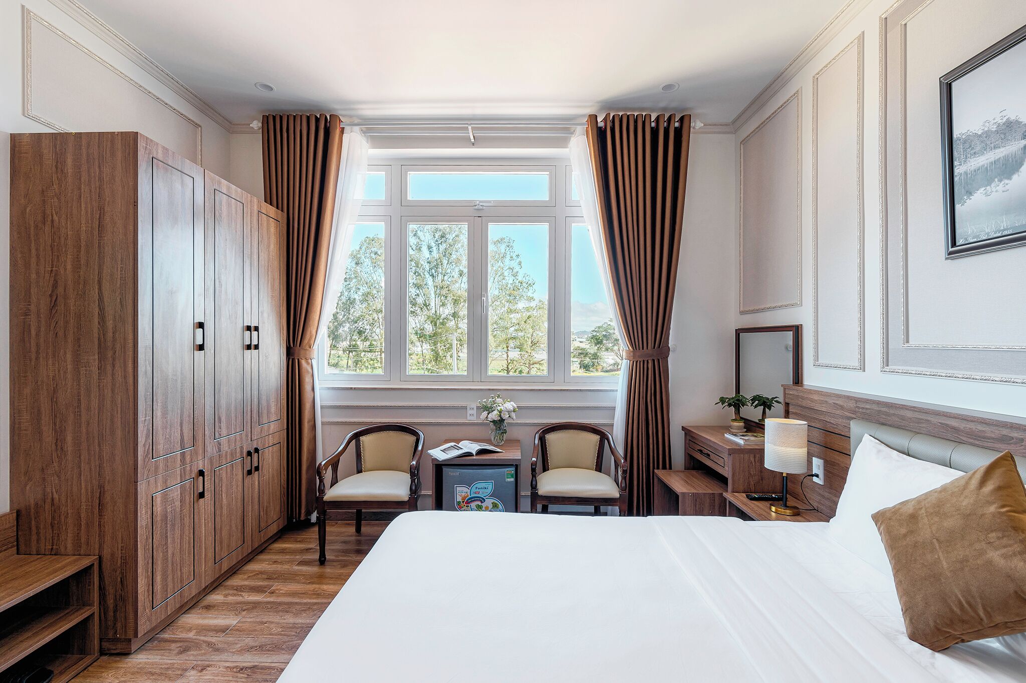 Une chambre d’hôtel avec un grand lit, deux chaises, un bureau avec une lampe, une petite table avec une plante et une armoire.