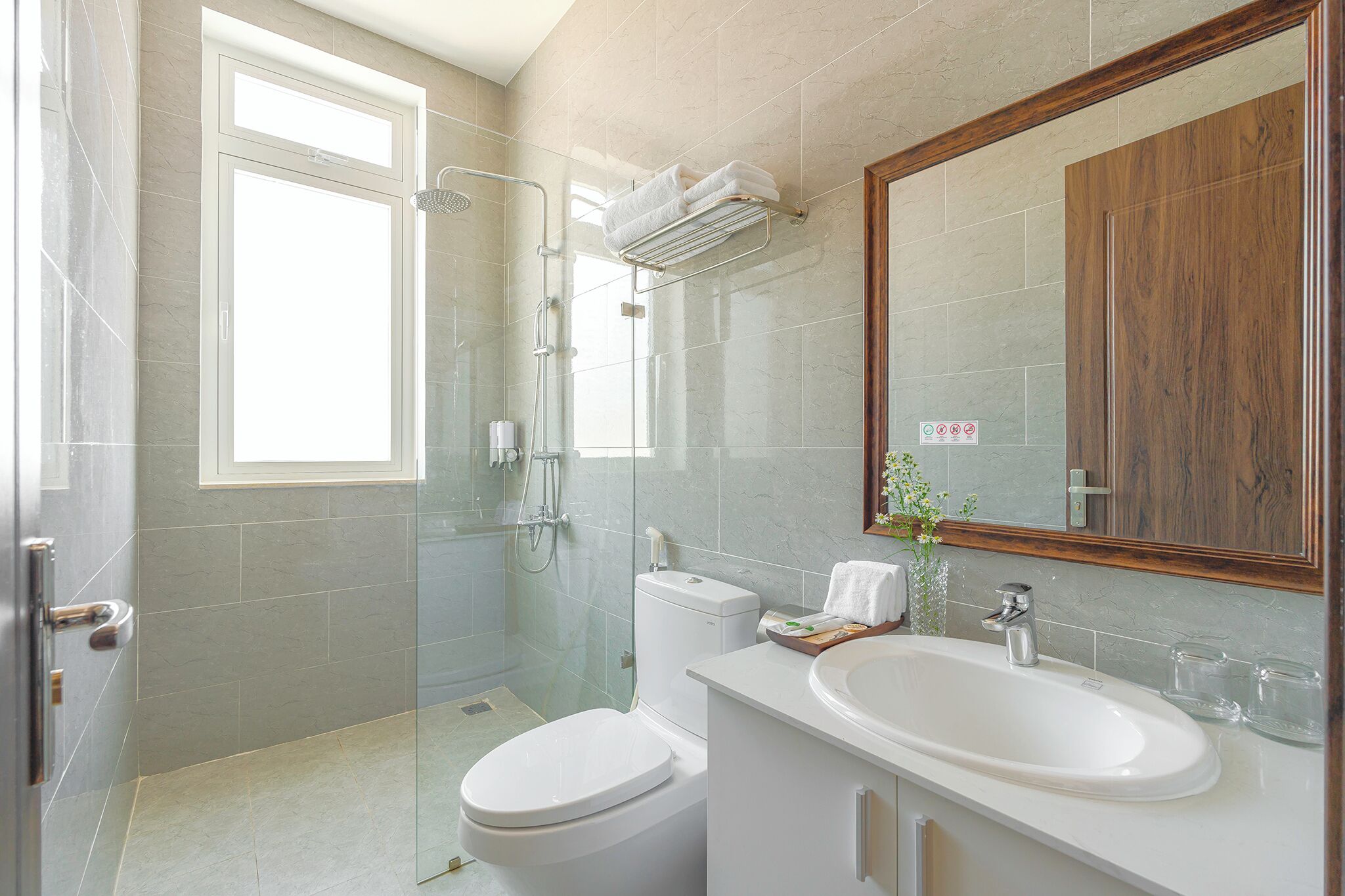 A modern bathroom with a glass shower enclosure, a toilet, a sink with a mirror, and a window providing natural light.