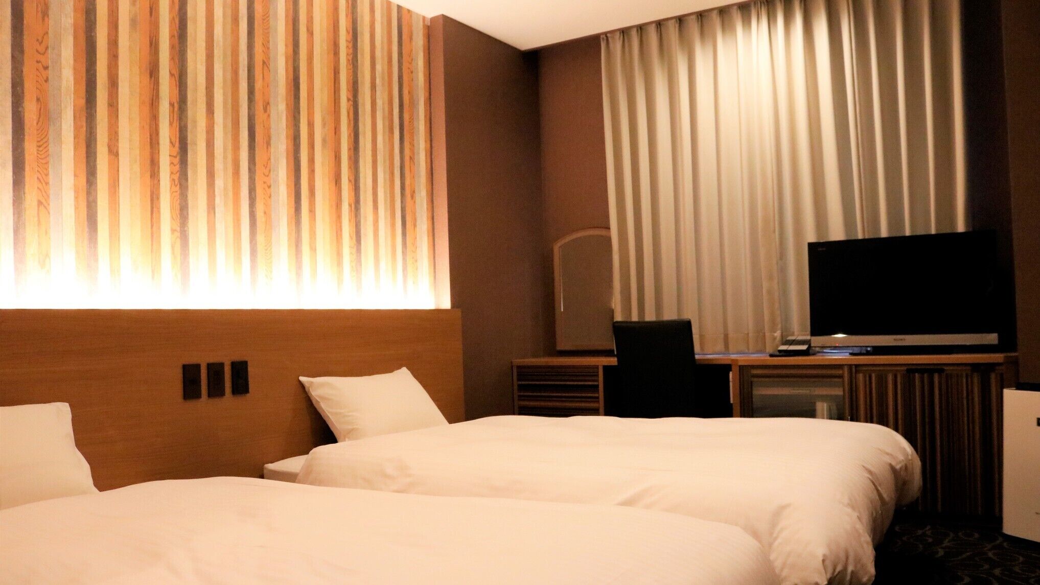 A hotel room with two beds, a TV, a desk with a computer, and a large window with curtains.