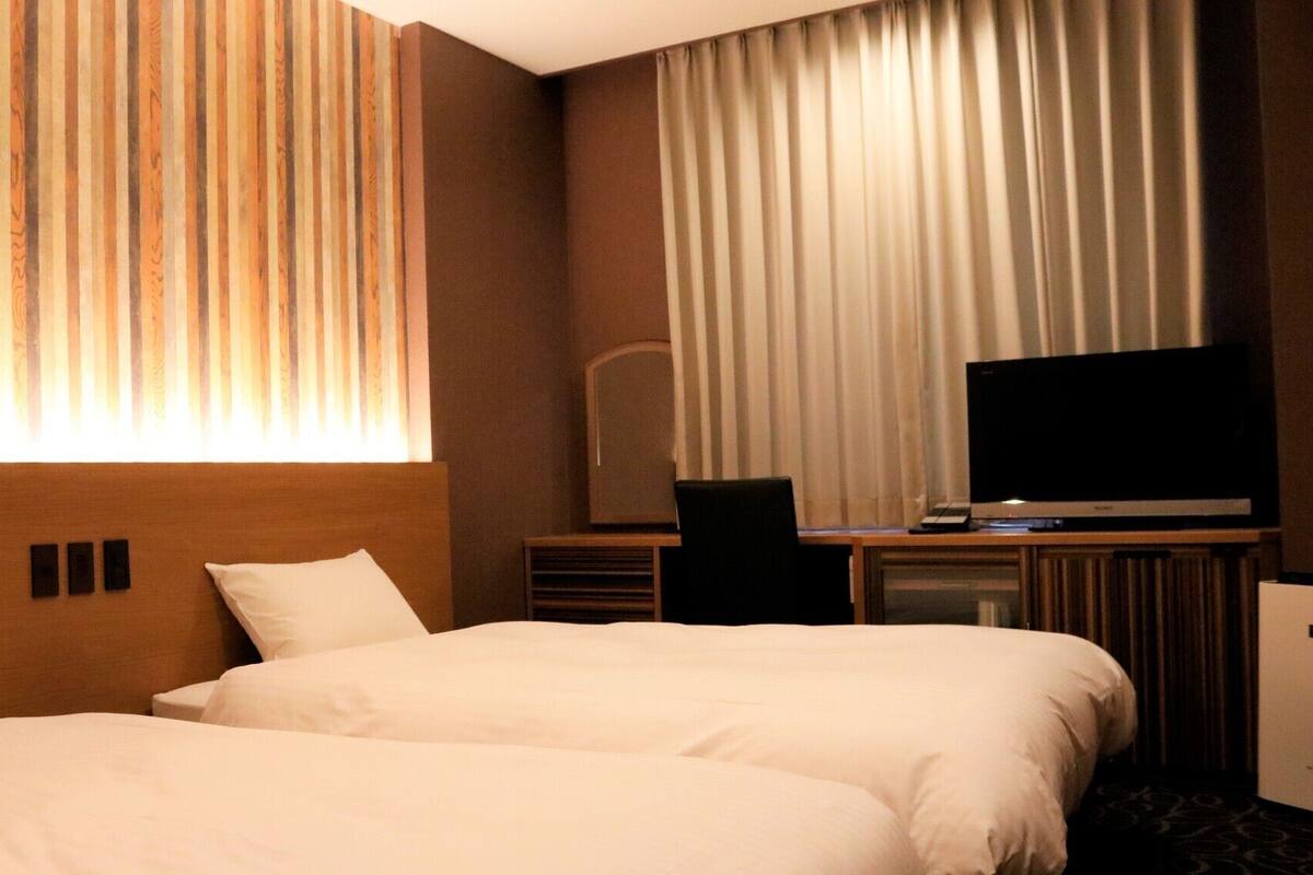 A hotel room with two beds, a TV, a desk with a computer, and a large window with curtains.