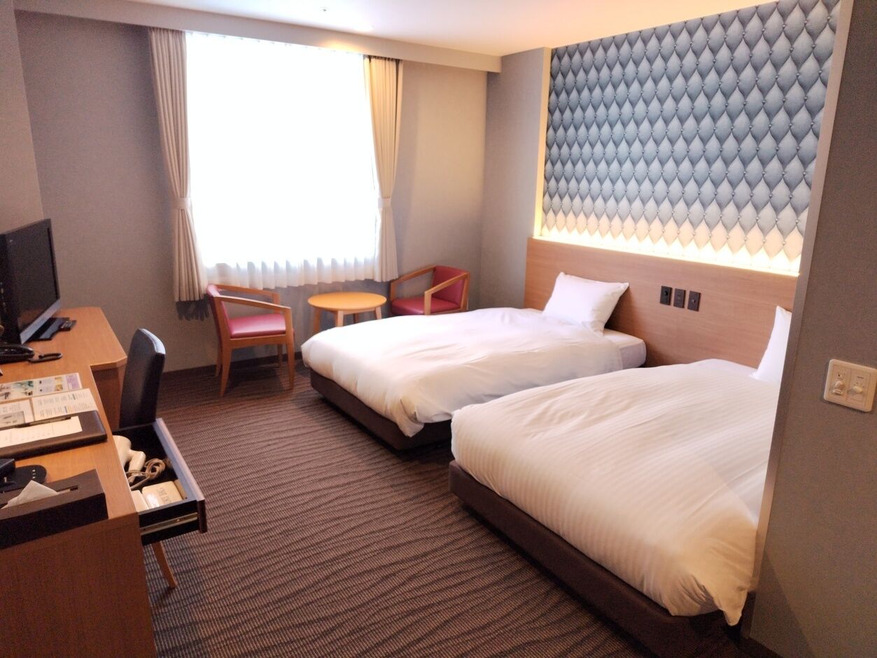 A hotel room with two beds, a desk, a chair, a TV, and a window with curtains.