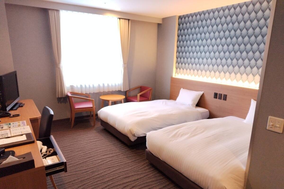 A hotel room with two beds, a desk, a chair, a TV, and a window with curtains.