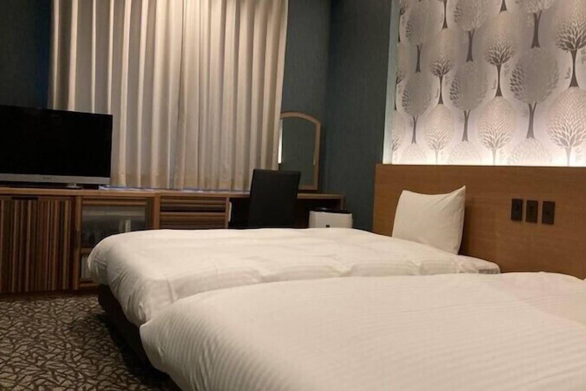 A hotel room with two beds, a TV, a desk, and a wall with a tree pattern.