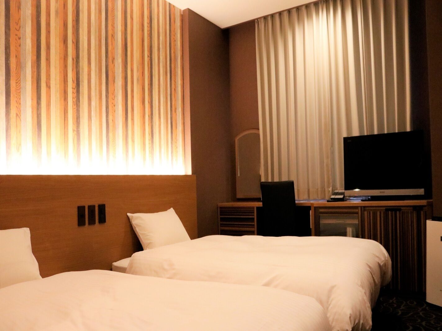 A hotel room with two beds, a television, a desk, and a large window with curtains.