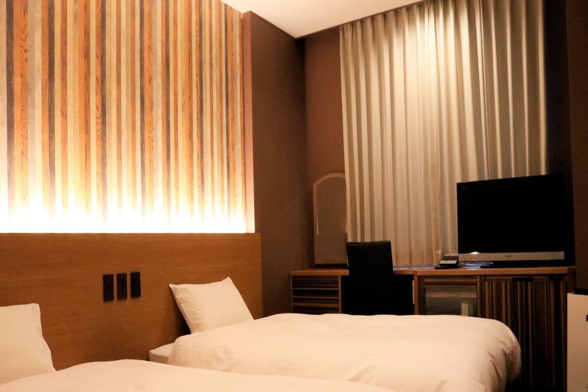 A hotel room with two beds, a television, a desk, and a large window with curtains.