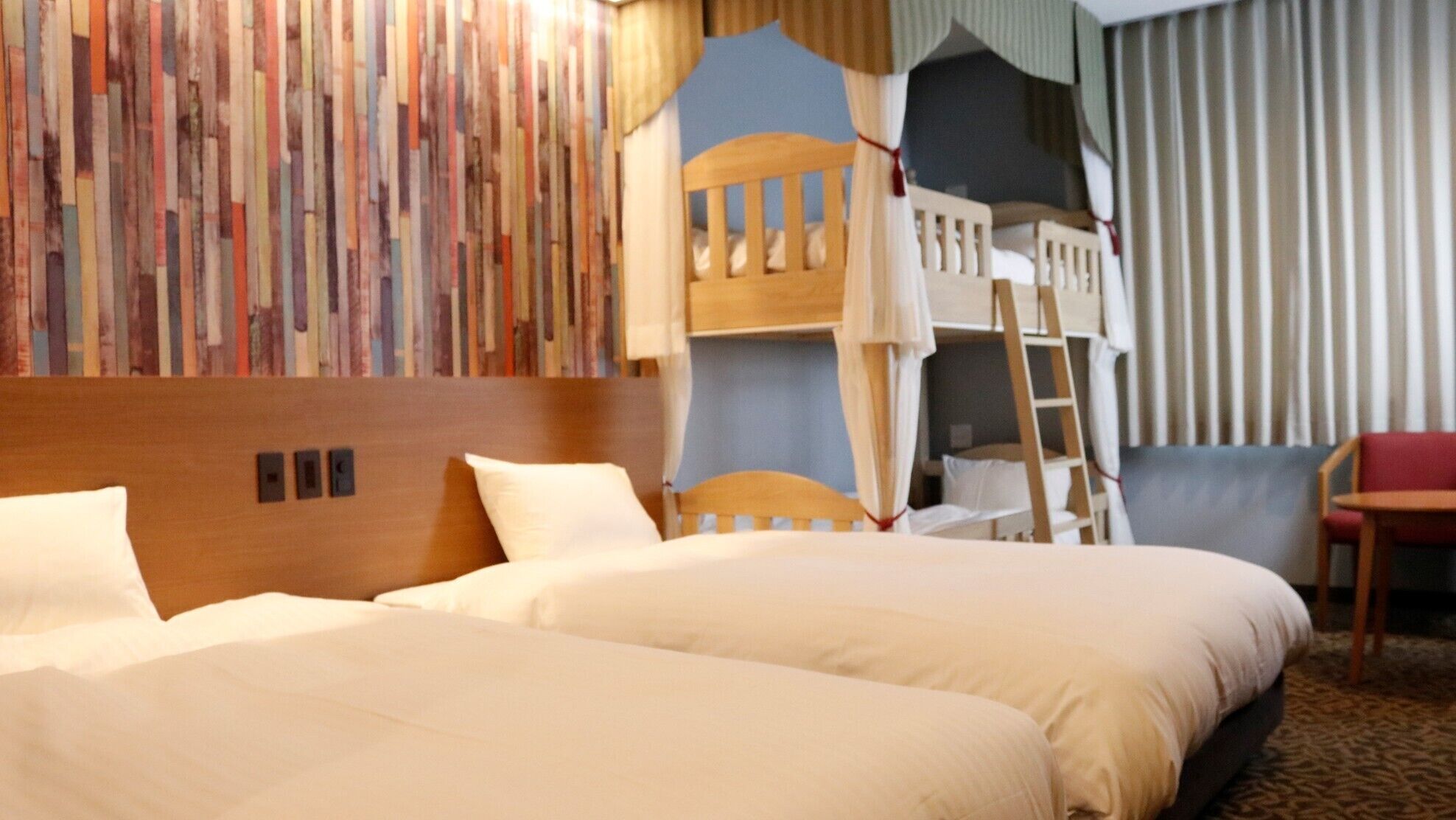 A hotel room with two beds, a bunk bed, a wooden headboard, a red chair, and a patterned wall.