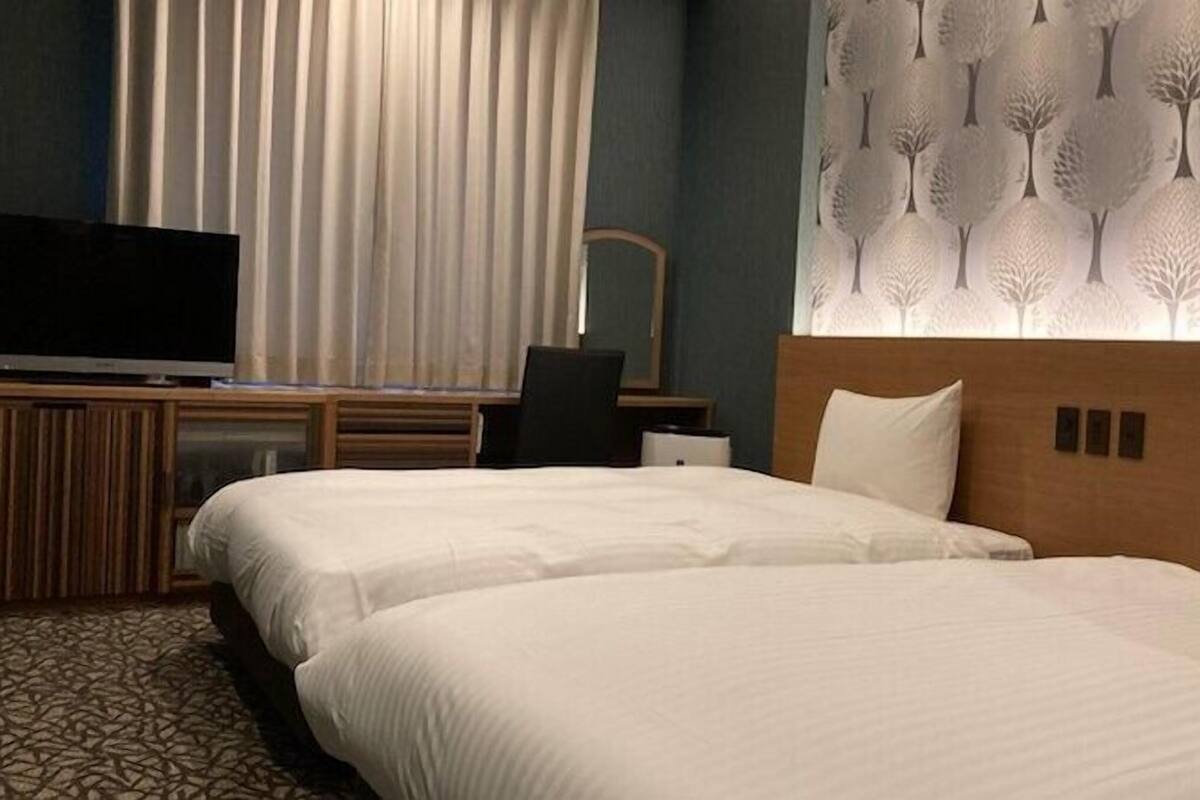A hotel room with two beds, a TV, a desk, and a wall with a tree pattern.